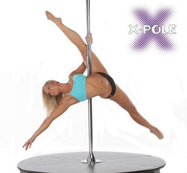 Blond woman wearing black shorts and aquamarine top, she is tanned, she is smiling and making a figure with open legs and holding only with one hand to the pole side ways, Her head is on the left side of the image and her split legs are on the right side of the image. She is Pole dancing on an x pole stage. White background, The image has the logo of X POLE.